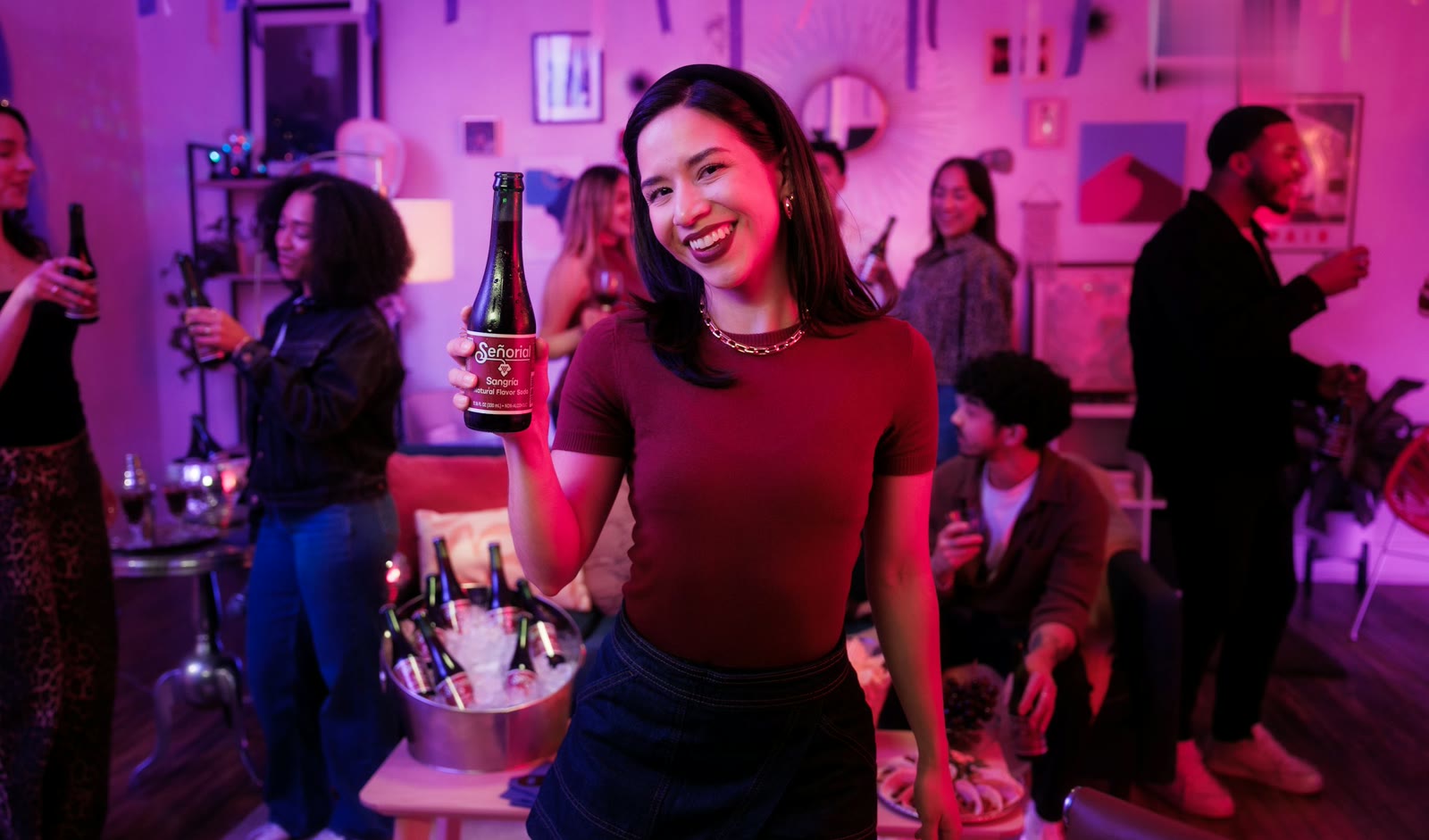 Woman smiling and raising a drink at a house party, friends chatting behind her