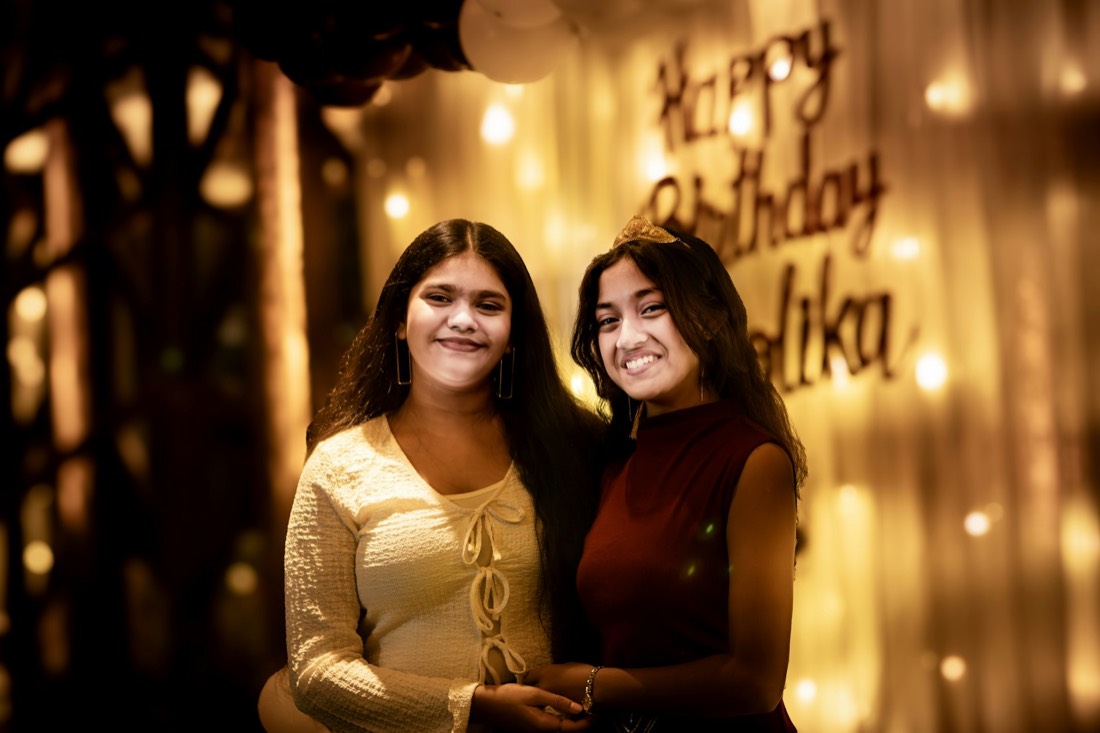 Two friends smiling in front of a Happy Birthday sign and string lights