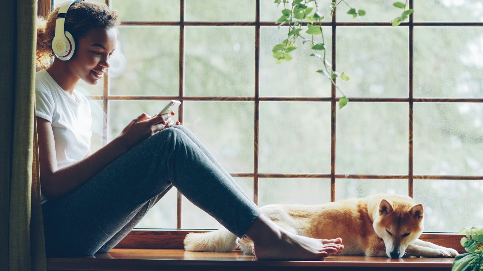 A woman with headphones listening to music on a sunny window sill at home, her dog resting beside her
