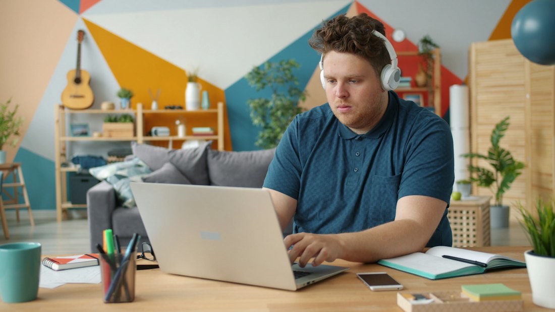 A man with headphones working on a laptop at a desk in a bright room